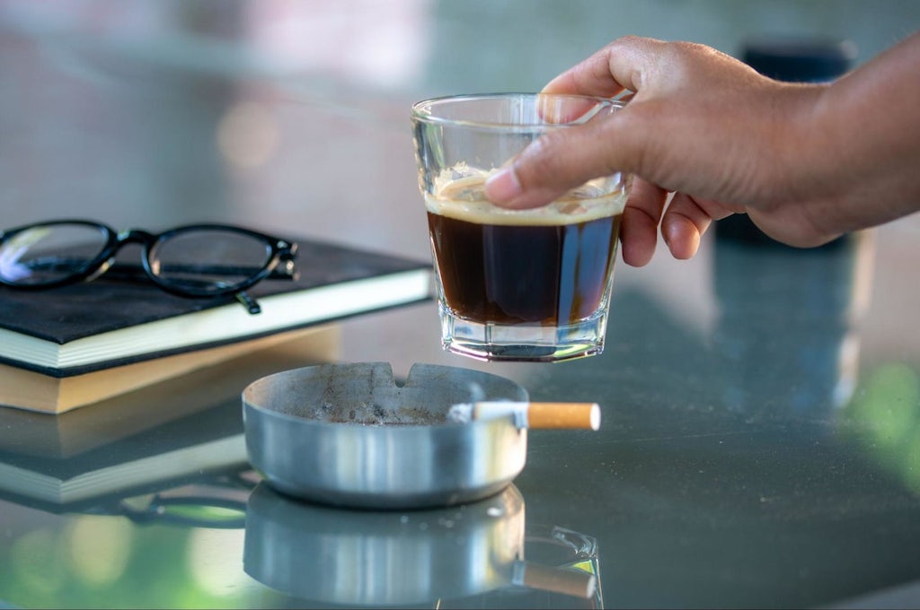 A hand holding a glass of black coffee and cigarette smoking on the table with the books and the glasses